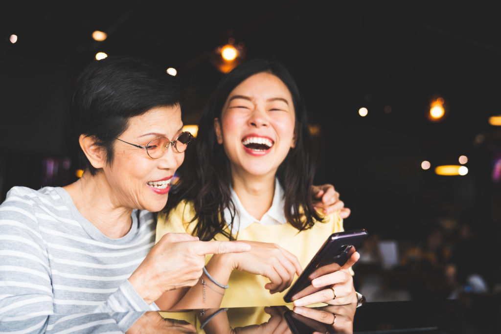 Asian mother and daughter laughing and using smartphone together at restaurant or cafe